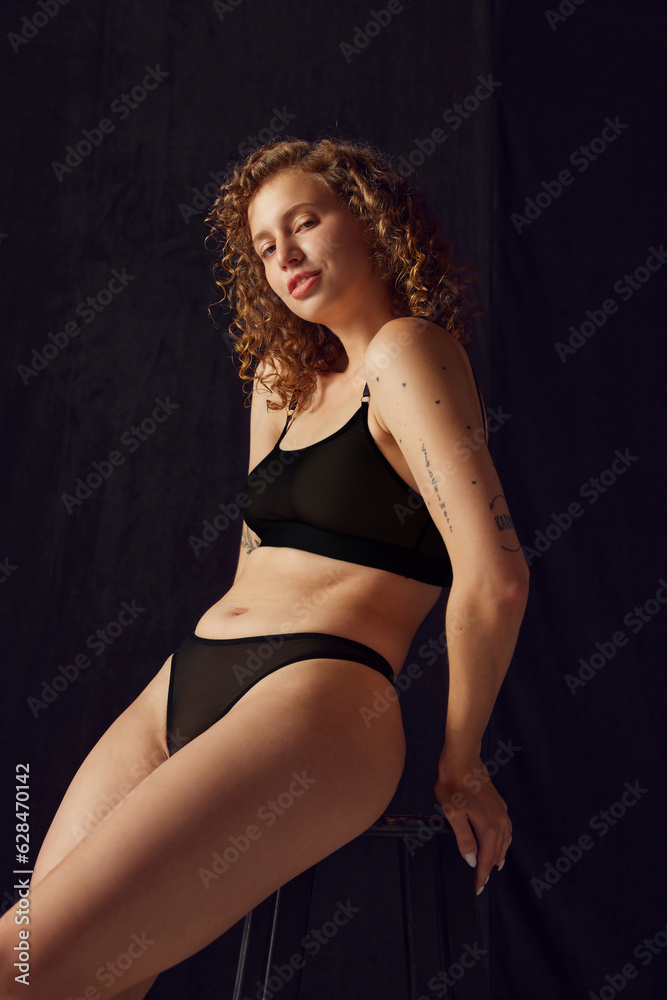 Fototapeta premium Portrait of fit woman in black underwear sitting on black chair and looking at camera on black studio background.