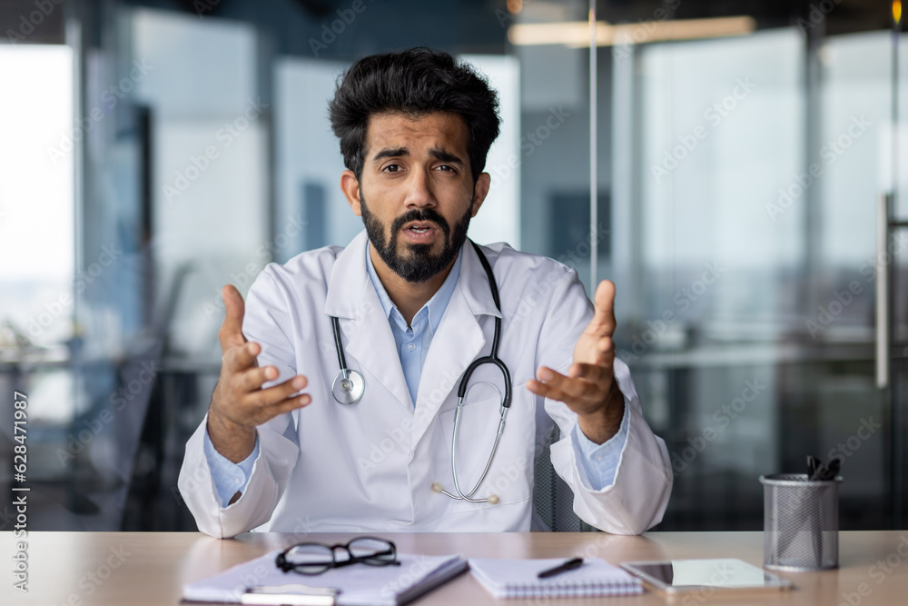 Portrait of a young Indian doctor talking seriously and worriedly to the camera. He sits in the office and communicates with the patient, conducts an online webinar, gestures with his hands