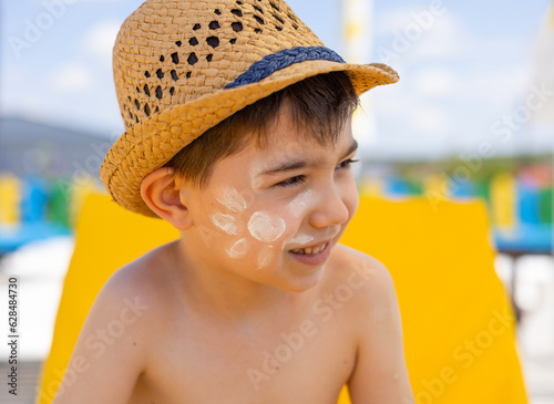 adorable preschooler kid boy with sun shape drawn from spf protection cream lotion on face cheek.child sitting on lounger,smiling,wearing beach hat summer vacation pool day.