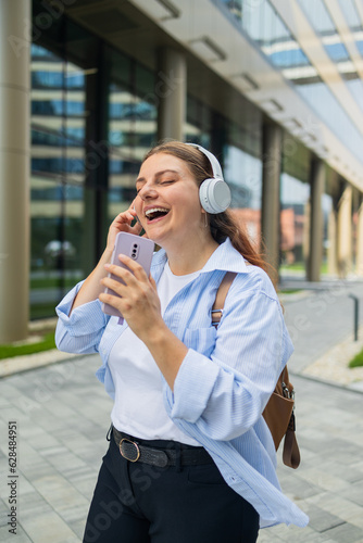 Young happy woman wearing suit using wireless earphones holding smartphone listening to music and singing, walking on urban city street. High quality photo © Shi 