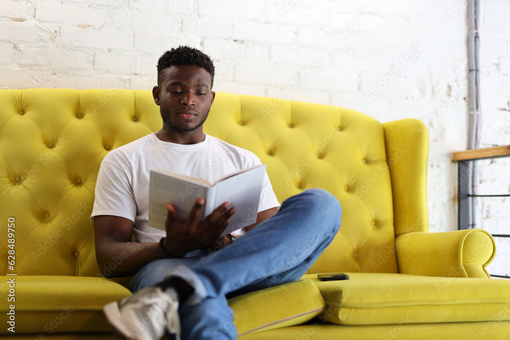 Confident African American man who is sitting on a yellow couch and reading a book. He is a young and casual student, business or literature who is studying.