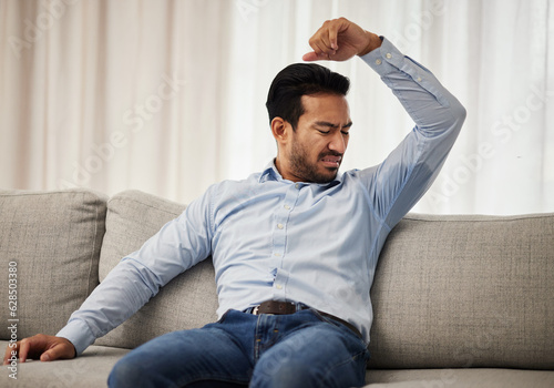 Armpit stink, smelling and a man on the sofa with a hygiene or bacteria problem. Bad, home and an Asian person with body odor, underarm sweat or grooming issue on the living room couch after work