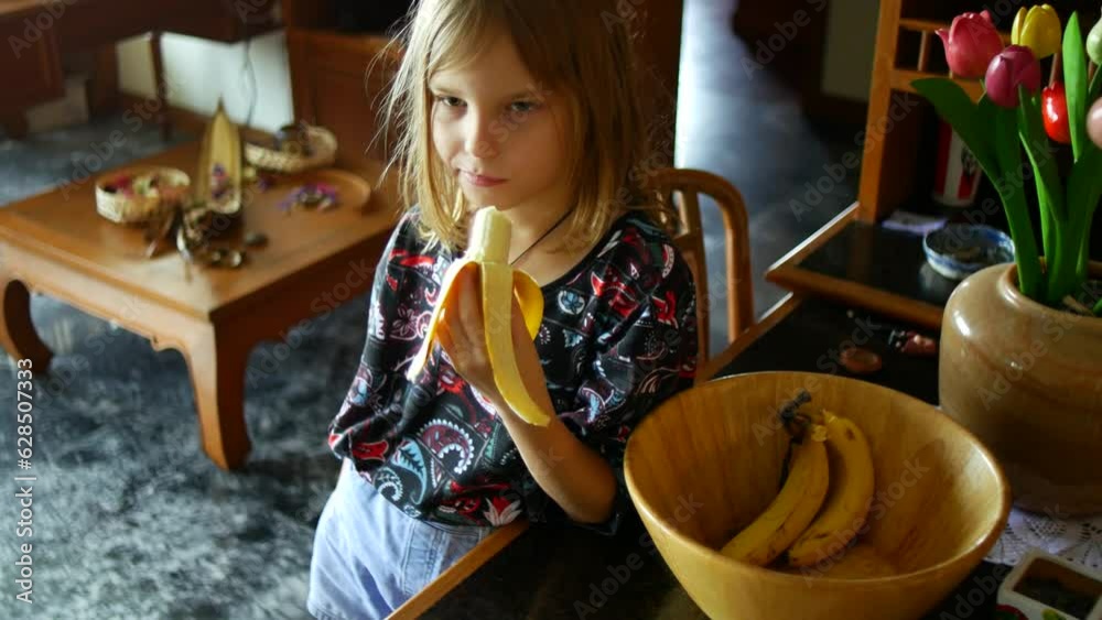 The girl eats a banana in the kitchen. Snack between online lessons.