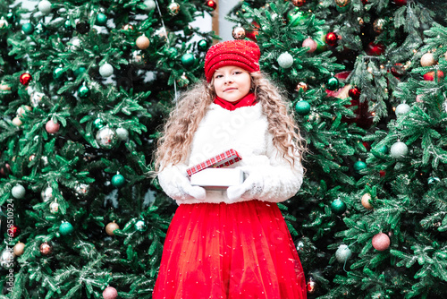 Gifts for Christmas and New Year, girl in a white fur coat and a red hat and skirt next to the Christmas Tree receives gifts outdoor, celebration and gifts photo