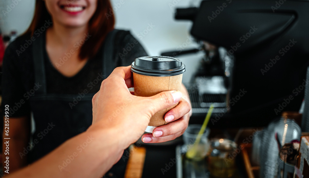 A friendly smiling female barista is holding in hands hot coffee in a takeaway paper cup submitted to the customer in a coffe shop. Small business concept