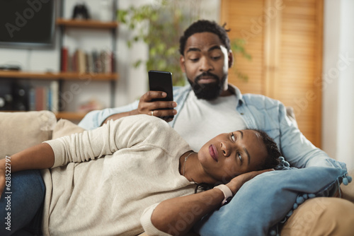Young black woman lying on her husband's lap and wondering to whom is he sending a text message while relaxing together on sofa