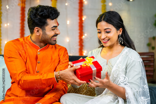 Happy young husband giving present to wife during festival celebration at home - brother giving gift to sister during raksha bandhan.