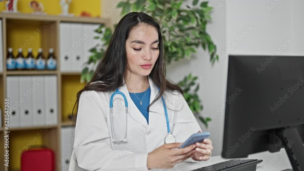 Young beautiful hispanic woman doctor using smartphone with serious face at the clinic Young beautiful hispanic woman doctor using smartphone with serious face at the clinic