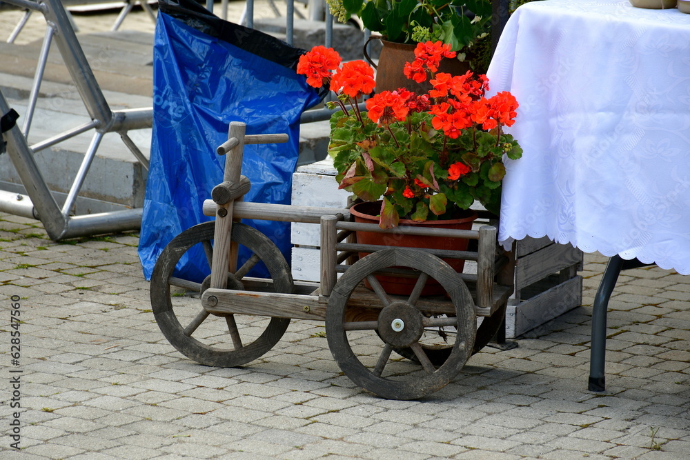 A close up on a wooden cart or trolley on three wheels used as a rural or rustic pot to grow plants and flowers in situated in the middle of a public square or public park next to some tables