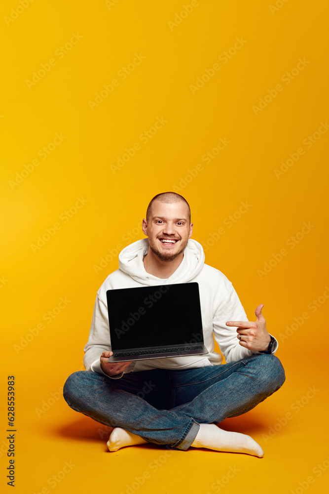 Vertical photo of hipster man wearing white hoodie smiling and pointing at new laptop in hands while sitting on floor, isolated over yellow background Vertical photo of hipster man wearing white hoodie smiling and pointing at new laptop in hands while sitting on floor, isolated over yellow background