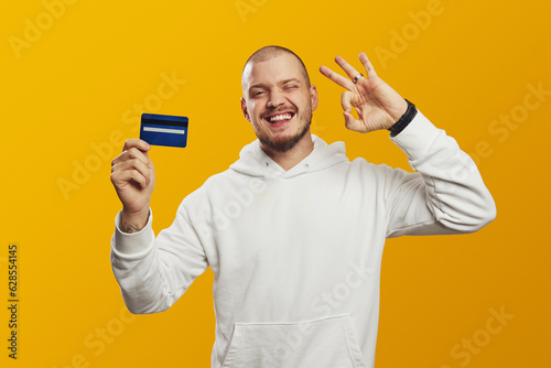 Excited young caucasian man in white hoodie holding credit bank card while showing OK gesture, blinks eye, isolated over yellow background
