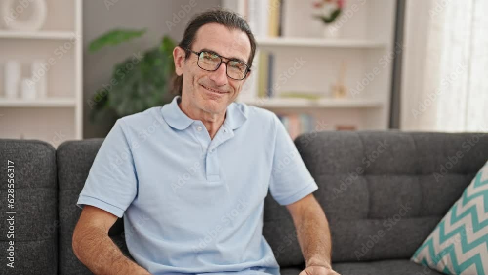 Middle age man wearing glasses sitting on sofa smiling at home Middle age man wearing glasses sitting on sofa smiling at home