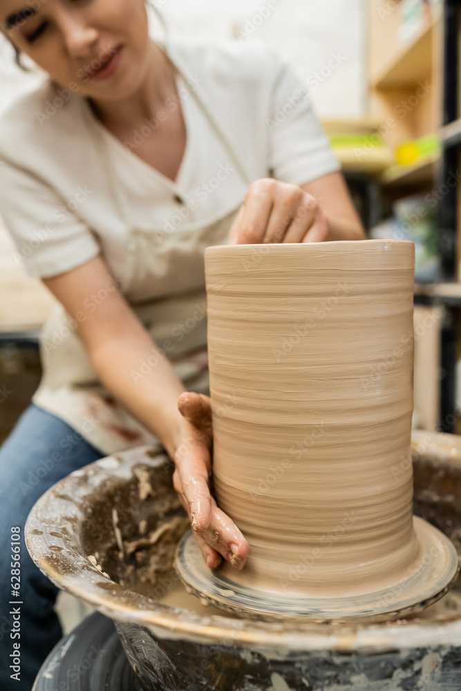 Cropped view of blurred female potter making clay vase on pottery wheel in workshop at background Cropped view of blurred female potter making clay vase on pottery wheel in workshop at background