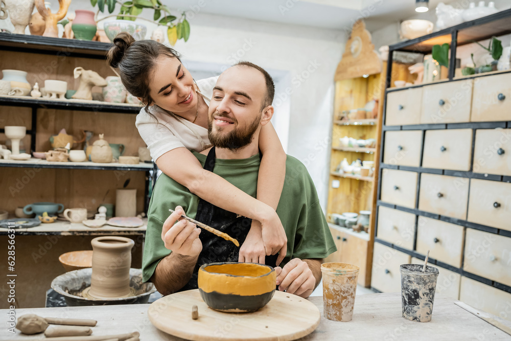 Smiling female potter hugging boyfriend coloring clay bowl while working in ceramic workshop Smiling female potter hugging boyfriend coloring clay bowl while working in ceramic workshop