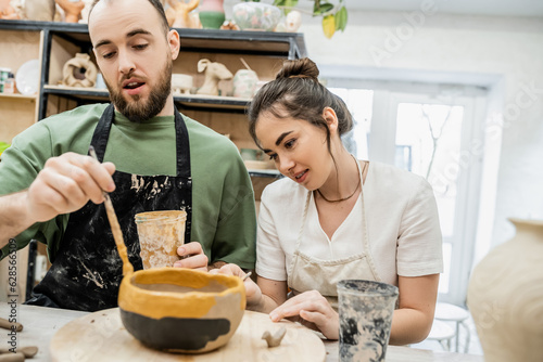 Smiling craftswoman talking to boyfriend while coloring clay bowl in ceramic workshop at background photo