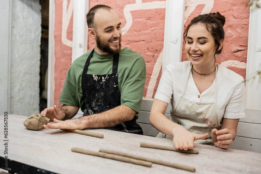 Joyful couple of sculptors in aprons modeling clay and talking in ceramic workshop Joyful couple of sculptors in aprons modeling clay and talking in ceramic workshop