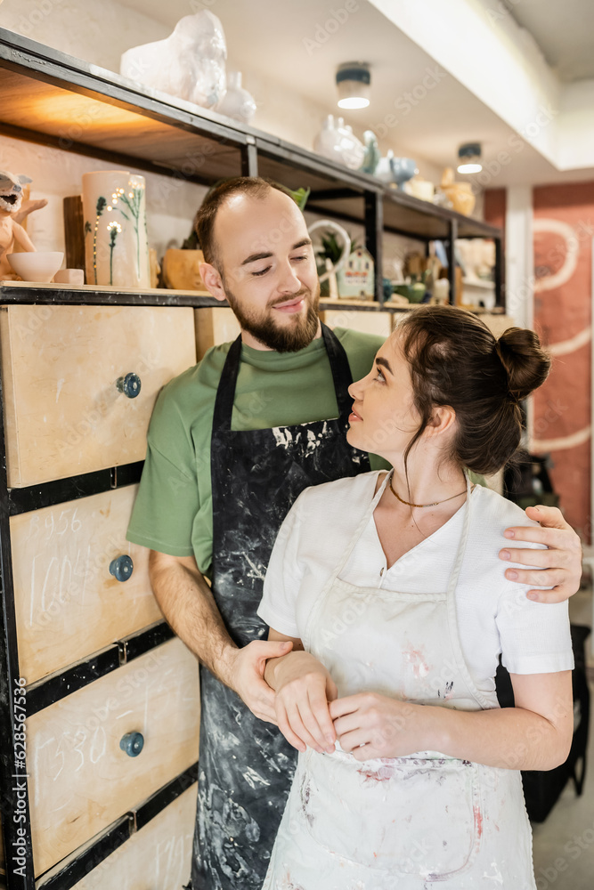 Couple of artisans in aprons smiling at each other while standing near rack in ceramic studio Couple of artisans in aprons smiling at each other while standing near rack in ceramic studio