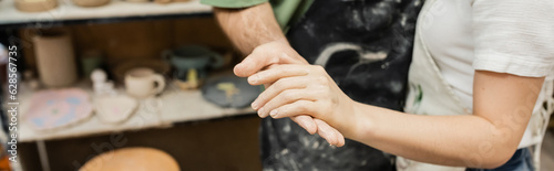 Cropped view of couple of potters in aprons holding hands while working in ceramic workshop, banner