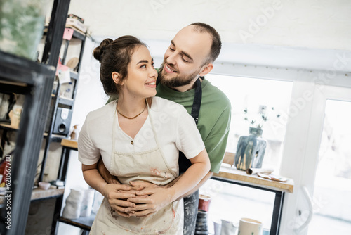 Smiling bearded artisan in apron hugging and looking at girlfriend in ceramic workshop