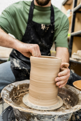 Partial view of blurred artisan in apron shaping clay on pottery wheel in ceramic workshop
