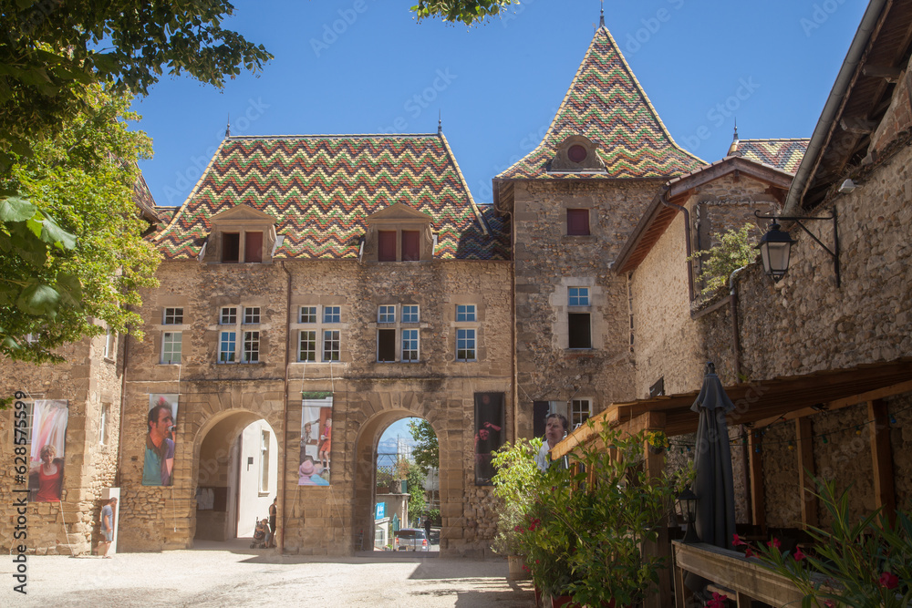 Ensemble architectural recouvert de tuiles vernissées et des arches accédant à une place du village de Saint-Antoine l'abbaye (Isère)