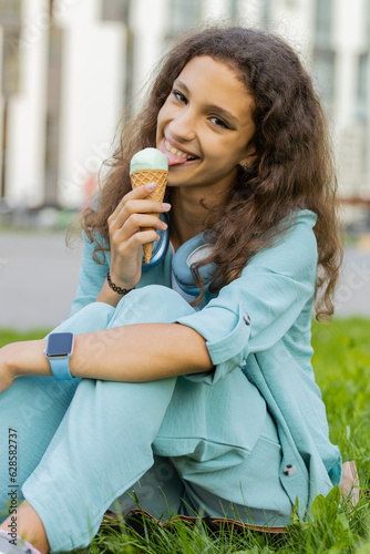 Happy pretty preteen child kid standing and eating ice cream in waffle cup. Cute smiling tween girl enjoys yummy ice cream licking  relaxing on summer holidays in urban european city street. Vertical
