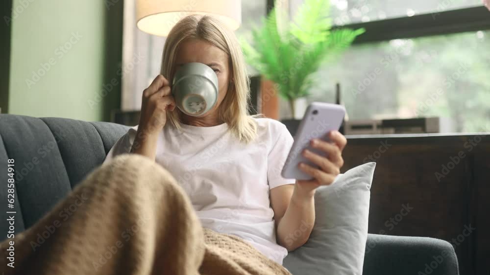 Portrait of charming blond mature woman with cup of hot coffee or tea hold smartphone scrolling watching social media indoors Happy female texting on her phone enjoying leisure time at home
