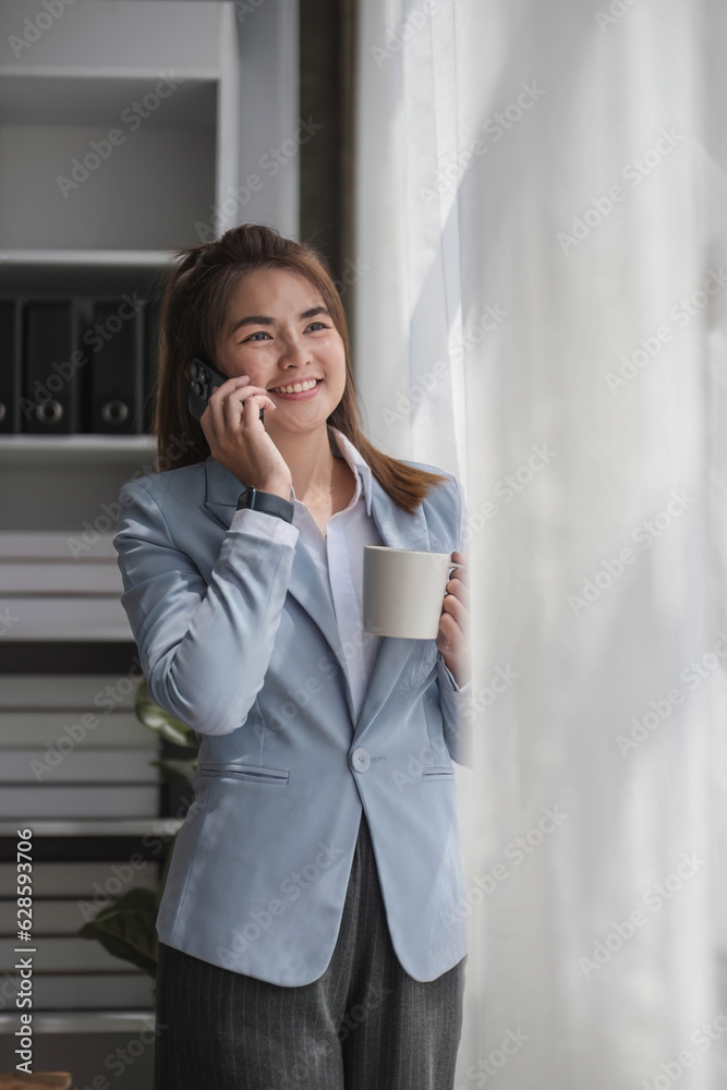 Portrait of young business woman with standing in office near the window and talking on mobile phone hand holding a cup of coffee Portrait of young business woman with standing in office near the window and talking on mobile phone hand holding a cup of coffee