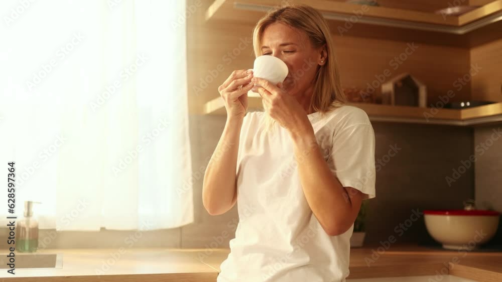 Portrait of beautiful mature woman looking out the window drinking hot drink of tea or coffee while the rays of the rising sun fall on her face at home kitchen Beautiful morning concept