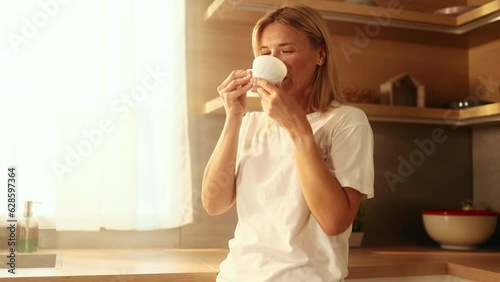 Portrait of beautiful mature woman looking out the window drinking hot drink of tea or coffee while the rays of the rising sun fall on her face at home kitchen Beautiful morning concept