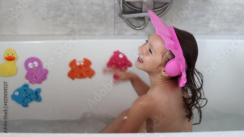 The cute little girl of European appearance giggles and splashes in his bath in slow motion, creating a spray of water. Water treatments. Pure happiness and carefree nature of childhood