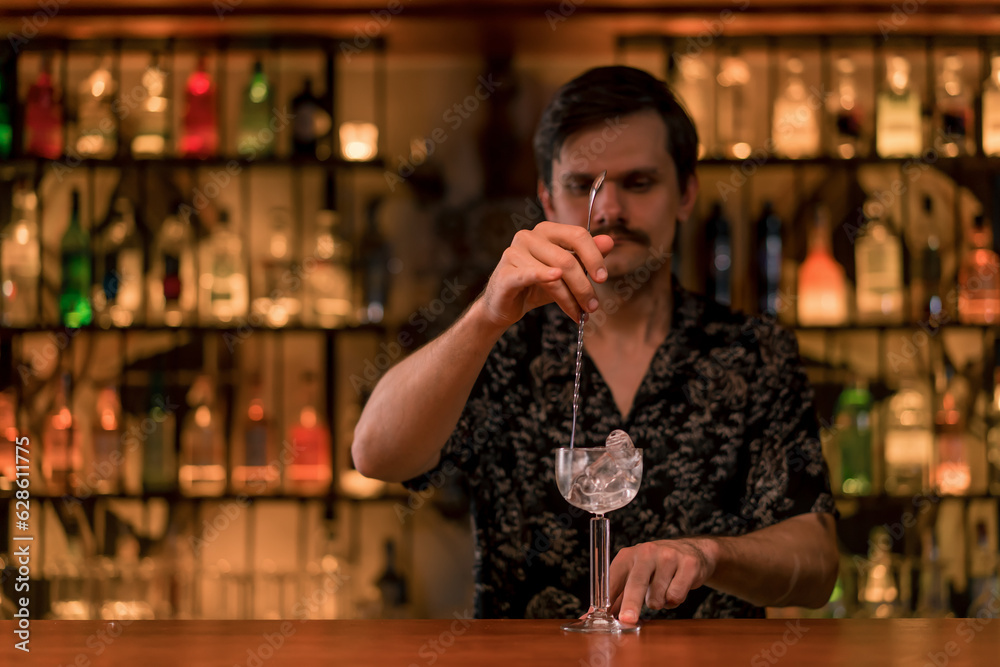 A bartender stirs ice with a cocktail spoon in a cocktail glass on the bar in a cocktail bar during the cocktail preparation process