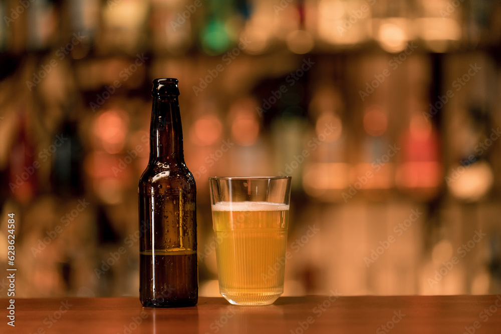 Close up of a bottle of beer standing next to a glass of beer on a bar counter in a club bar on a blurred background