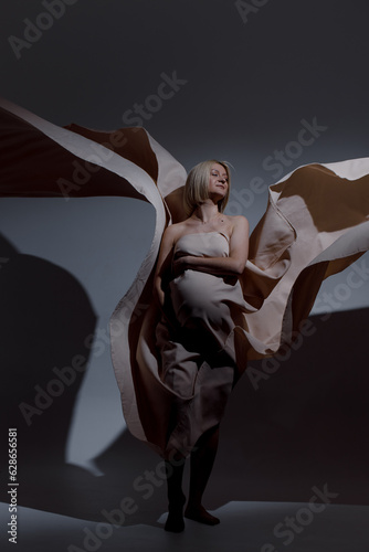 A pregnant woman with white hair of Caucasian appearance is wearing a beige sheet that flies beautifully on the background.