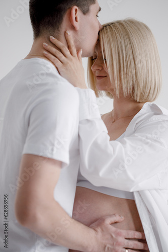 Family couple. A pregnant blond woman of Caucasian appearance is in bed with her husband, who kisses her on the forehead and gently touches her belly. They wear jeans and a white T-shirt.
