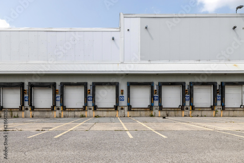 High bay warehouse facade showing long row of empty numbered truck loading bays, rubber door seals, target lights, empty asphalt parking lot in foreground, blue sky with clouds, nobody