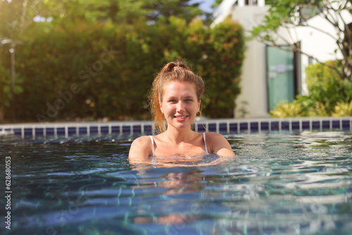 Portrait of happy cheerful young woman having fun, swim in swimming pool outdoors at summer sunny day, enjoy summer vacation, look at camera 