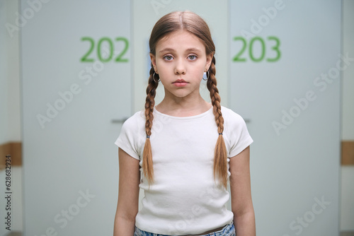 Sad child with pigtails stands in a bright hospital corridor photo