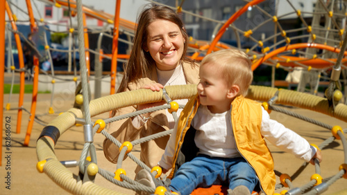 Portrait of happy smiling mother swinging her baby son on the playground on sunny day. Children playing outdoor, kids outside, summer holiday and vacation.
