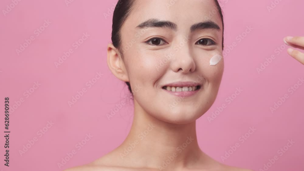 Portrait of a young smiling beautiful asian woman with pure skin applying the moisturising cream on her cheek on pink background. Concept of benefits of cosmetics in skin care Portrait of a young smiling beautiful asian woman with pure skin applying the moisturising cream on her cheek on pink background. Concept of benefits of cosmetics in skin care