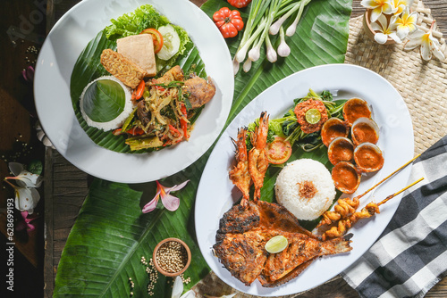 Balinese cuisine of seafood contains baked fish, clamp, and prawn served with another amin course of tempe and tahu with indonesian dressing vegetables on the table covered with banana leaf. photo