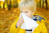 Little boy sneezing and wipes nose with napkin during walking in autumn park
