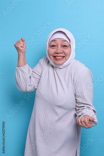 Elderly Asian Muslim woman 60s celebrate clenching fists isolated on blue background. 