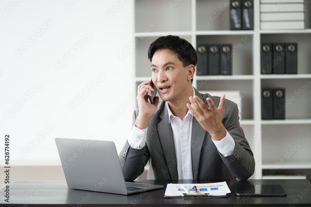 Young Asian businessman having a phone call with his client while sitting in the office room. Young Asian businessman having a phone call with his client while sitting in the office room.