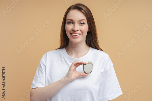 Canvas Print Young smiling woman holding cardio stimulator, pacemaker looking at camera isolated on beige background