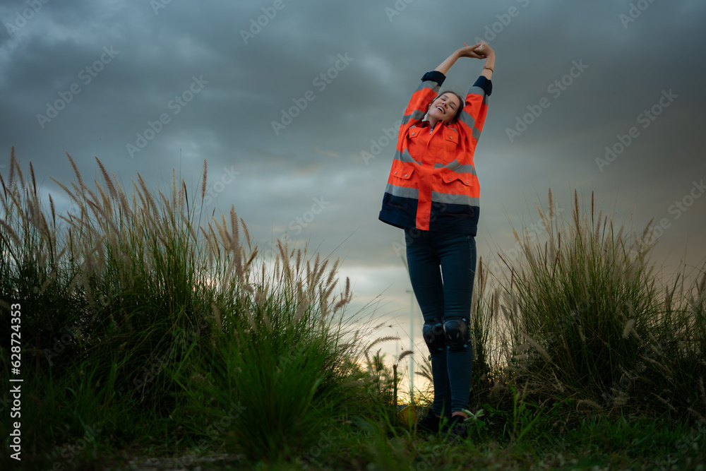 Young woman in engineer uniform and high visibility with raised arms standing on grassy field at sunset, The concept of relax time after work