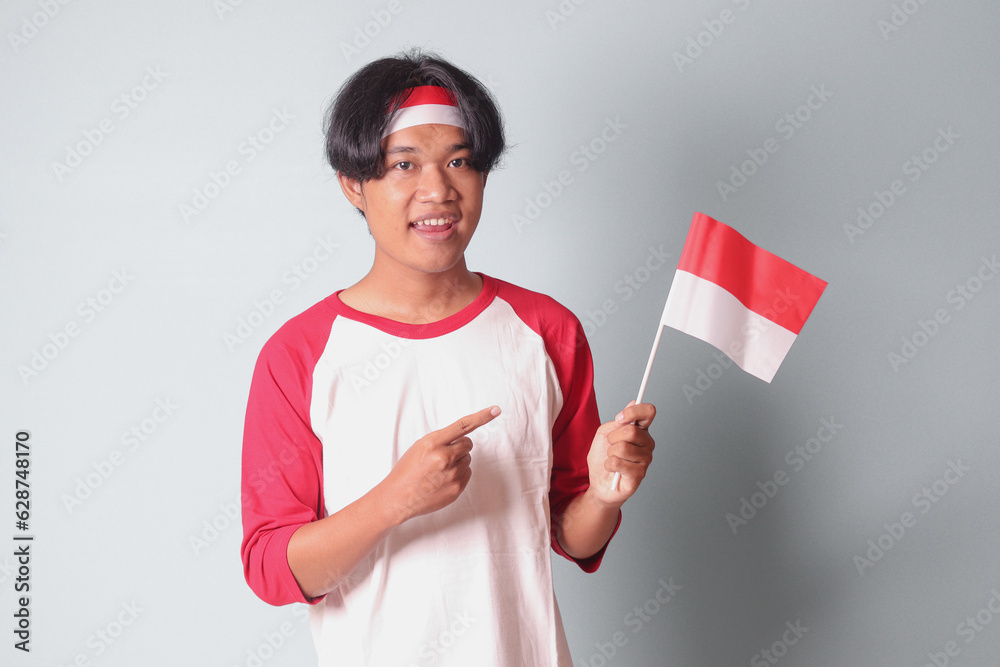 Portrait of attractive Asian man in t-shirt with red and white ribbon on head, showing product and poiting away while holding indonesian flag. Isolated image on gray background Portrait of attractive Asian man in t-shirt with red and white ribbon on head, showing product and poiting away while holding indonesian flag. Isolated image on gray background