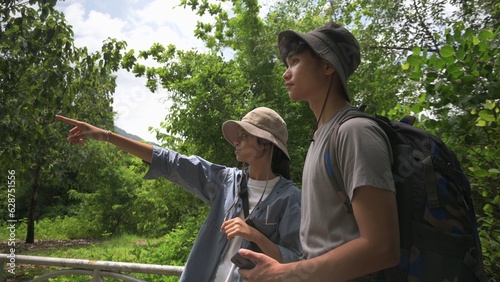 Two Asian young teen backpackers looking for directions while hiking in tropical forest park in summer holiday. Outdoor pursuits. Teenager hobbies and leisure activities.