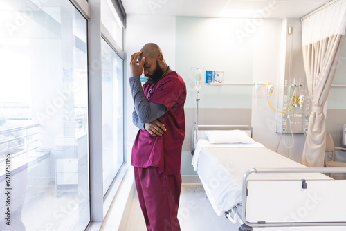 Sad african american male doctor wearing scrubs looking through window at hospital