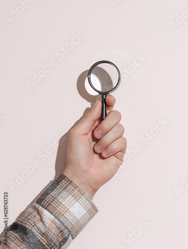 Man's hand in shirt holding magnifying glass on beige pastel background with shadow